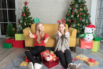 Obraz na płótnie Canvas Two beautiful caucasian girls sit on the living room floor in front of Christmas tree, wear an antler hat and thumbs up, look at camera with a happy smiling face, Christmas and Happy New Year concept