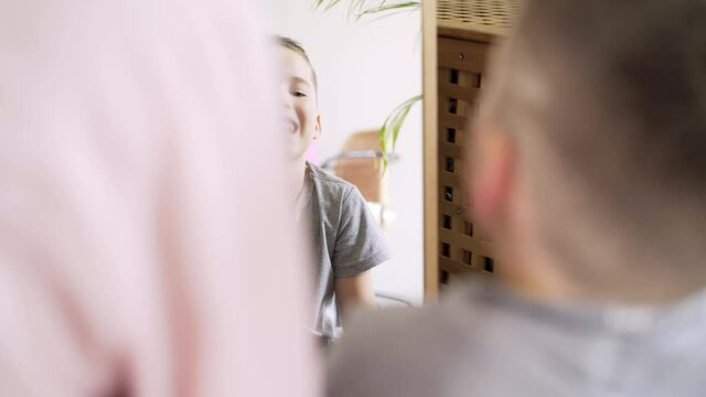 cute little boy in grey t-shirt does logopedic exercises with tongue sitting on floor near large mirror in light living room with family close view