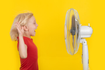 Fair-haired child standing in front of fan and is happy. Bright yellow background. Strong heat.
