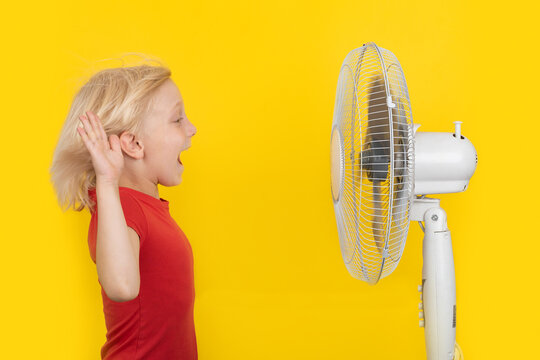 Fair-haired Child Standing In Front Of Fan And Is Happy. Bright Yellow Background. Strong Heat.