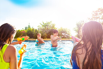 Portrait with two teen girls in swimming pool shooting water-gun squirt pistol in boys on sunny day