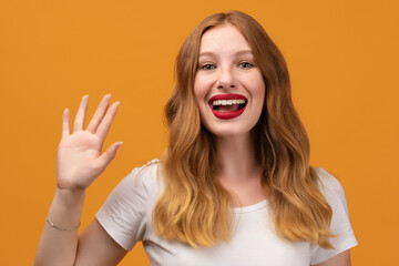 Portrait of friendly woman with wavy redhead, standing waving hand, looking at camera with engaging toothy smile