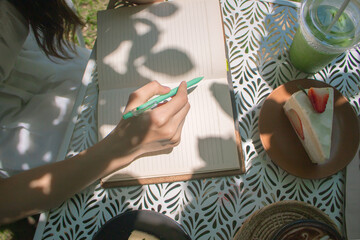 Women hands are writing on notebook in the garden at cafe. Relaxation and reading concept. Weekend activity concept. Space for short list.