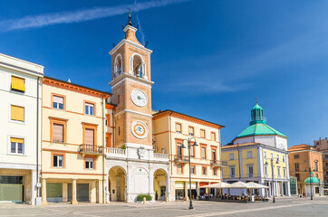 Fototapeta na wymiar Piazza Tre Martiri Three Martyrs square with traditional buildings with clock and bell tower in old historical touristic city centre Rimini with blue sky background, Emilia-Romagna, Italy