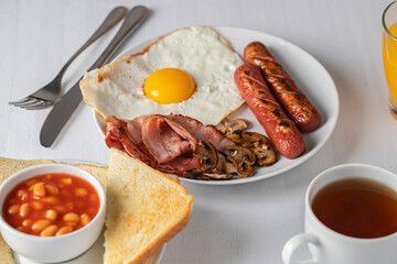 Typical English breakfast with fried eggs, sausages, bacon, beans, mushrooms, orange juice, toasts and tea on the table. Copy space