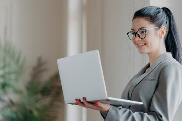 Indoor shot of busy corporate worker poses in coworking space, holds modern laptop computer, transmits date, connected to wireless internet, wears glasses for visual correction, sends feedback