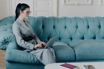 Obraz premium Content businesswoman works remotely in cozy home office, poses with laptop computer on sofa, wears formal elegant clothes, keyboards and searches necessary information in internet, sends emails