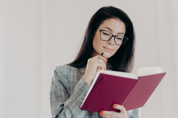 Fototapeta premium Serious brunette female management consultant uses notepad for planning organization, holds pen and writes down notes or list to do, wears formal clothing, has serious expression, looks in notebook