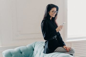 European entrepreneur wears corporate suit and spectacles, uses online banking application for transferring money, holds cellular, sends email, poses at back of comfortable sofa, uses free wifi