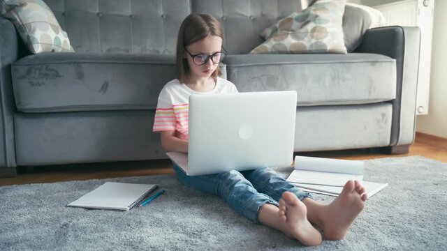 Serious little 8 years old girl in glasses sits on a carpet in a living room and typing on a laptop. Remote education technologies and homework.
