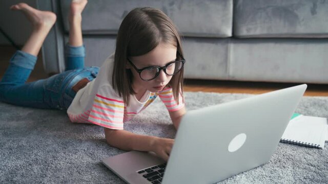 Serious little 8 years old girl in glasses lying on a carpet in a living room and typing on a laptop. Remote education technologies and homework.