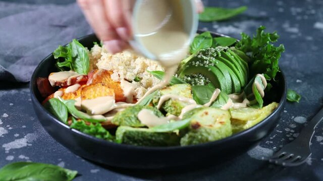 Vegan lunch plate. Quinoa salad with baked zucchini, sweet potato, avocado and tahini dressing in black plate, dark background.