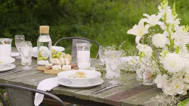 vintage wooden table with elegant festive setting and burning candles decorated with fresh white aromatic flowers stands in summer garden closeup