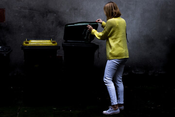 A girl in a yellow sweater throws garbage in a garbage can.Woman is throwing away the beer bottles.Woman is sorting the garbage.A young woman is putting a bag of rubbish in a large bin.Save the planet