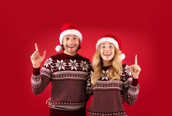 Joyful millennial couple in woolen sweaters and Santa hats pointing upwards on red background