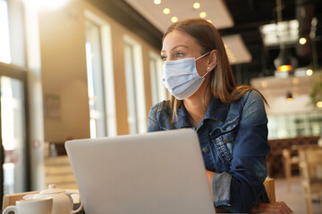 Young woman sitting at coffee shop and working on laptop with face mask