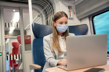 Businesswoman commuting by train, working on laptop and wearing face mask