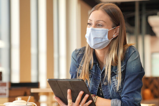 Young Woman Sitting At Coffee Shop And Working On Digital Tablet With Face Mask