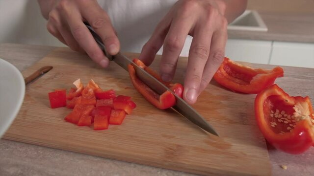 A guy in a white T-shirt cuts a red pepper with a sharp knife for making a salad. Light wood cutting board.
