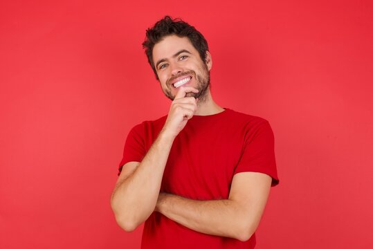 Young handsome caucasian man wearing t-shirt over isolated red background, Optimistic keeps hands partly crossed and hand under chin, looks at camera with pleasure. Happy emotions concept.