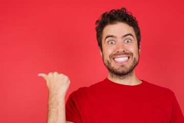 Lovely Young handsome caucasian man wearing t-shirt over isolated red background pointing aside with forefinger, showing at copy space having news about bargains