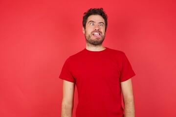 Young handsome caucasian man wearing t-shirt over isolated red background showing grimace face crossing her eyes and showing tongue . Being funny and crazy