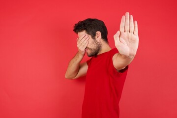 Young handsome caucasian man wearing t-shirt over isolated red background covers eyes with palm and...
