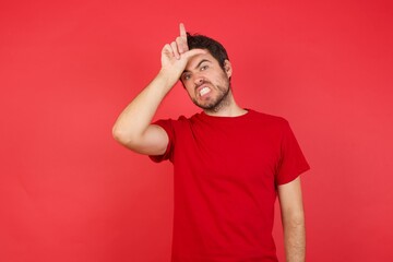 Fototapeta premium Young handsome caucasian man wearing t-shirt over isolated red background making fun of people with fingers on forehead doing loser gesture mocking and insulting.