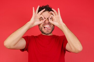 Young handsome caucasian man wearing t-shirt over isolated red background doing ok gesture like binoculars sticking tongue out, eyes looking through fingers. Crazy expression.