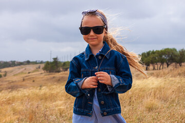 Adorable little girl in denim jacket, black sunglasses, blue plaid dress in yellow grass field. Happy stylish long blonde hair child on countryside landscape. Cute kid walking outdoor rural road trip.