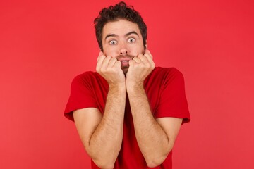 Photo of fearful Young handsome caucasian man wearing t-shirt over isolated red background keeps hands near mouth, feels frightened and scared,  has a phobia,  Shock and frighted concept.