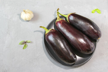 Fresh raw purple eggplants on a ceramic black plate with purple and green basil on stone background. Top view, copy space.