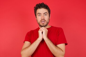 Young handsome caucasian man wearing t-shirt over isolated red background praying for luck has hands crossed near face, amazed and opening mouth looking front.