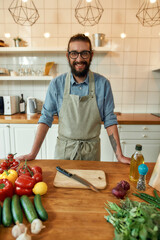 Young man, Italian cook in apron smiling at camera, ready for preparing healthy meal with vegetables in the kitchen. Cooking at home concept