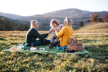 Small girl with mother and grandmother having picnic in nature at sunset.