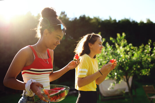 Side view of young teenager girls friends outdoors in garden, eating watermelon.