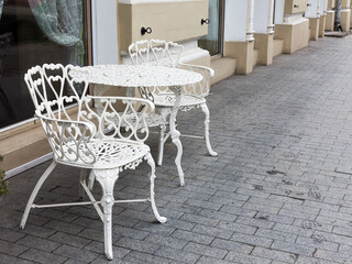 A vintage white forged table and two armchairs stand on the sidewalk