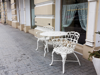 A vintage white forged table and two armchairs stand on the sidewalk