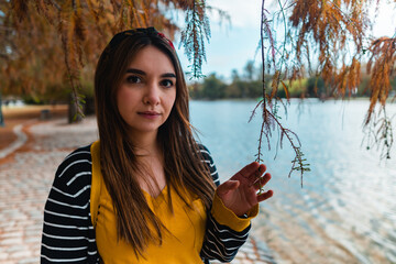 A young woman at autumnal park beside a lake looking at camera while touching a branch