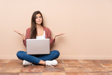 Teenager student girl sitting on the floor with a laptop having doubts while raising hands