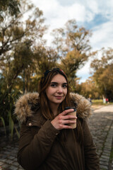 A pretty caucasian woman smiling while holding a coffee and enjoying a walk at park. Vertical