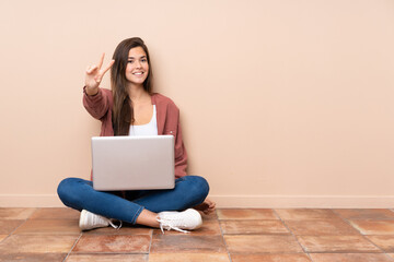 Teenager student girl sitting on the floor with a laptop smiling and showing victory sign