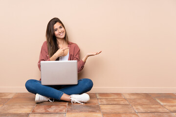 Teenager student girl sitting on the floor with a laptop holding copyspace imaginary on the palm to...