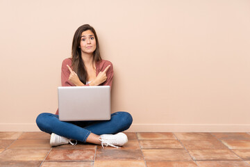 Teenager student girl sitting on the floor with a laptop pointing to the laterals having doubts