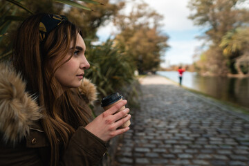 A pretty caucasian woman looking at park lake while holding a cup of coffee. Copy space