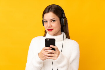 Teenager girl isolated on yellow background listening music with a mobile and looking front