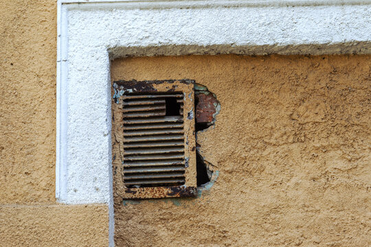 Old metal cooling grill on a textured building wall, colorful stains. The layers of paint exfoliate, entangled cracks and spots on the surface. Dark holes for ventilation