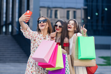 A group of young beautiful girls in casual clothes with sunglasses, makeup, hair hoop and colored shopping bags making selfie after successful shopping