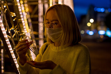 Fototapeta na wymiar Portrait of a young woman in a yellow sweater with a coronavirus mask, against the background of yellow city lights at night. Bokeh in the background. Woman with sanitizer gel. Coronavirus concept.