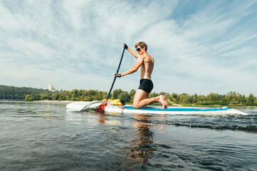 Athletic guy in sunglasses and a muscular body moves on the water on the sup board, actively rowing. Young man paddles on sup board on a sunny day